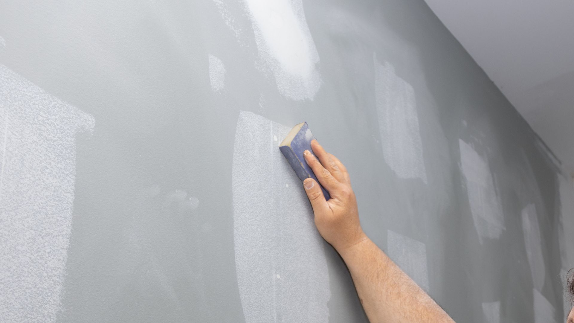 A Caucasian man hand using sanding block to smooth drywall patch.