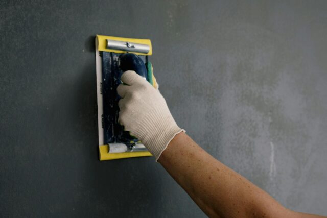 A gloved painter using sanding block on dark gray black wall.