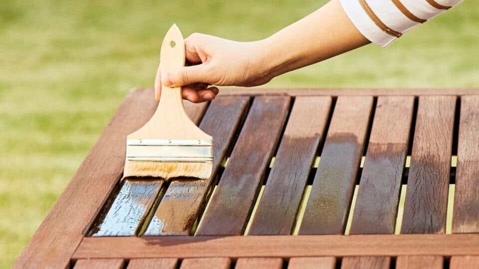 A hand holding a brush applying dark brown stain to wooden table outdoors.