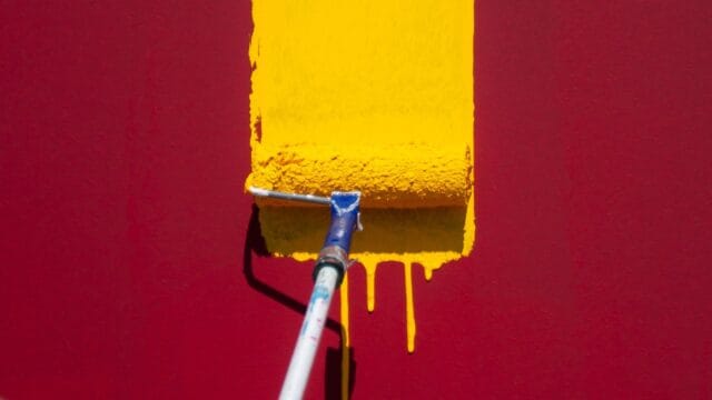 A paint roller dripping yellow paint down maroon red wall.