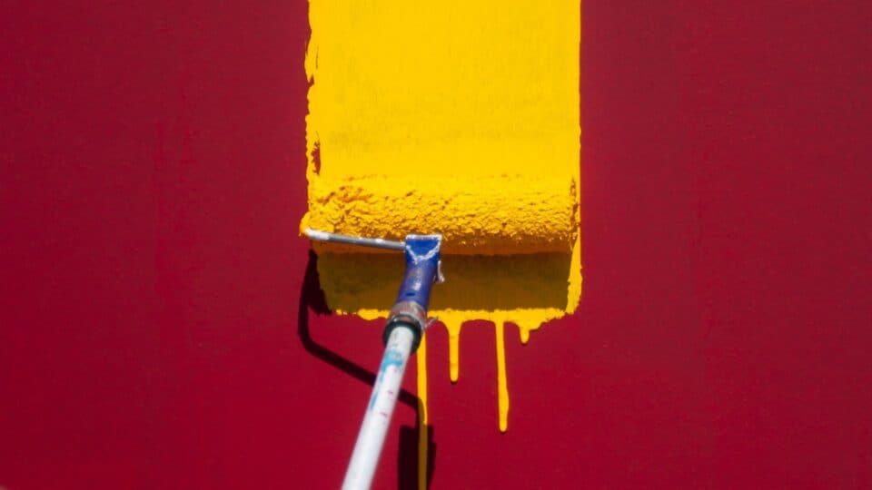 A paint roller dripping yellow paint down maroon red wall.