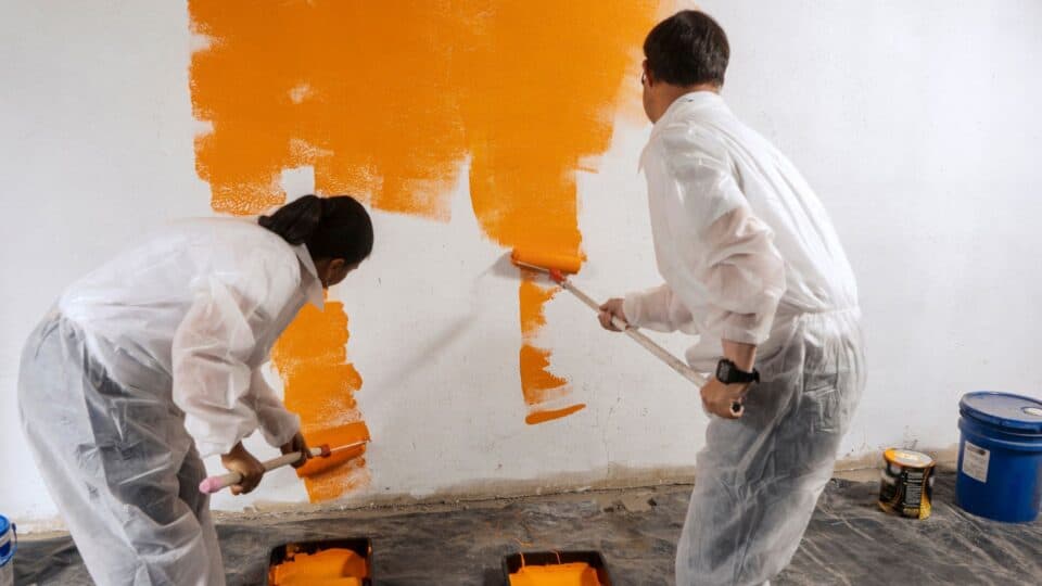 A young man and woman pair of painters painting over a white wall with a vibrant orange color.